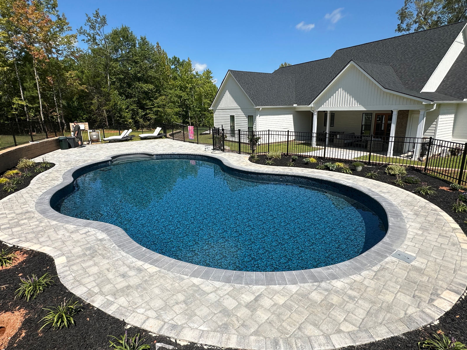 Freeform pool with a sweeping paver deck beside a modern farmhouse